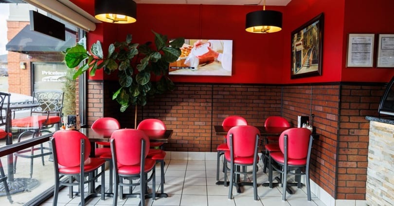 Restaurant interior with red walls, brick wainscoting, red chairs, dark tables, and a TV showing food.