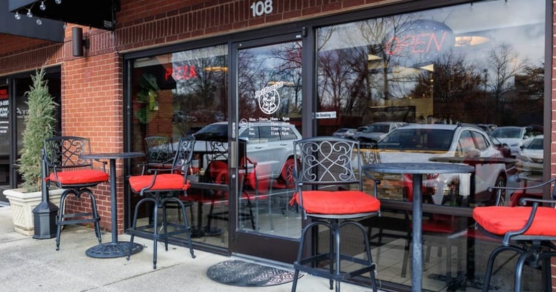Outdoor patio seating with black metal tables and chairs with red cushions in front of a brick building.